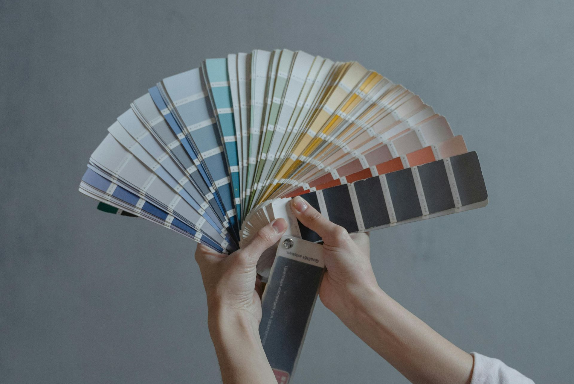 Hands holding a fan of paint color swatches against a gray wall. Colors range from blues to tans and blacks.