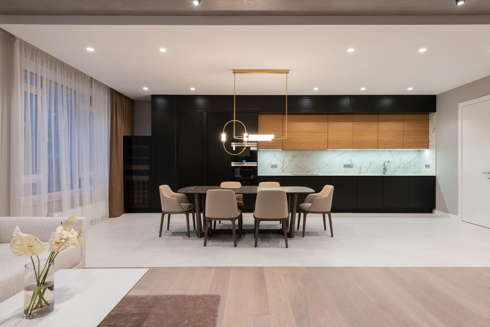 Modern dining room with a kitchen. Black and wood cabinets, table with chairs, and a decorative light fixture.