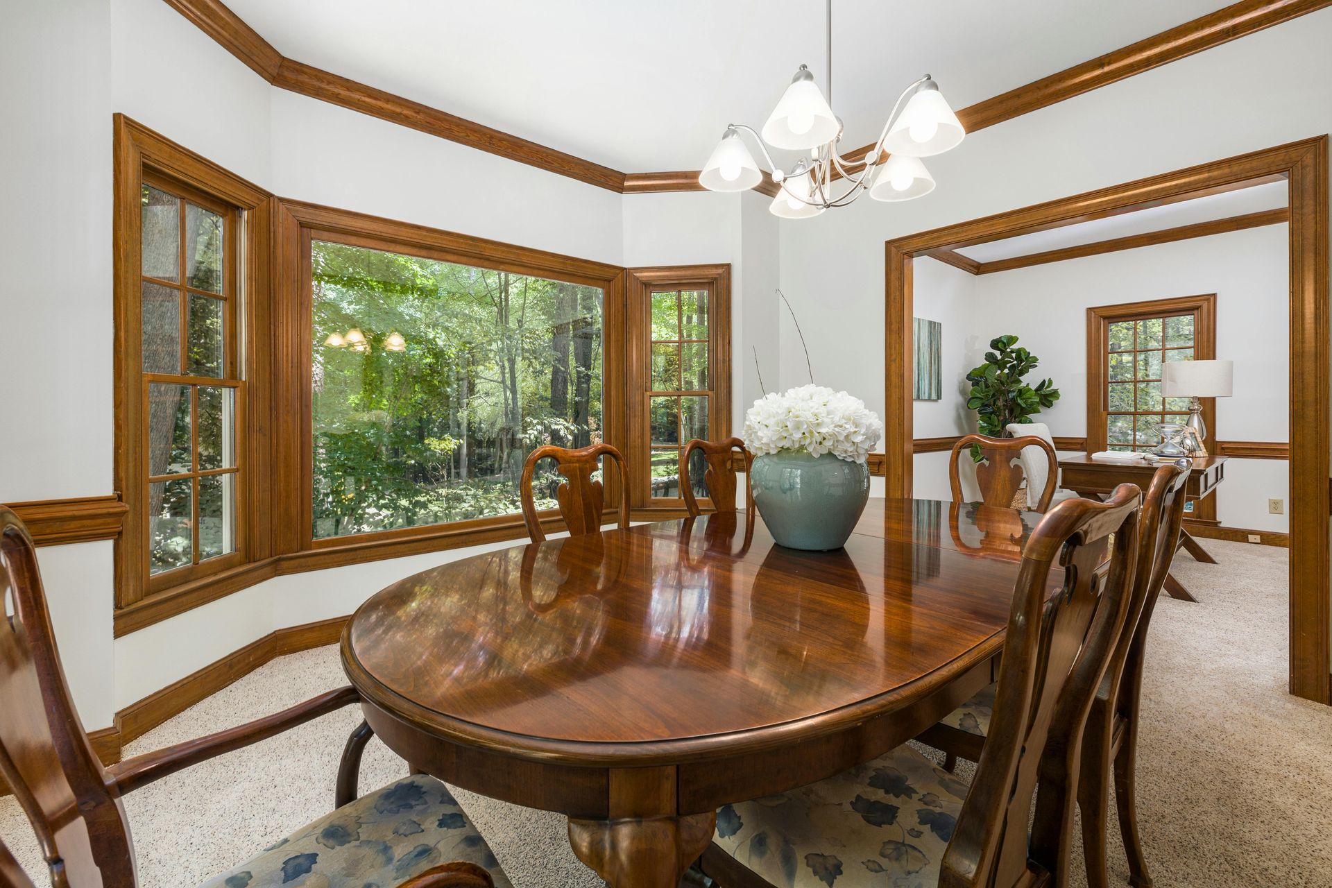 Dining room with oval wooden table, surrounded by chairs, large windows, and a chandelier.
