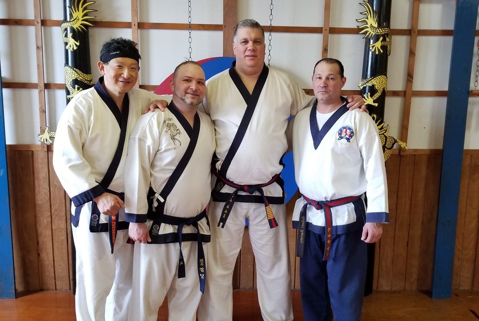 Four martial artists in white uniforms with black or red trim pose in a dojo.