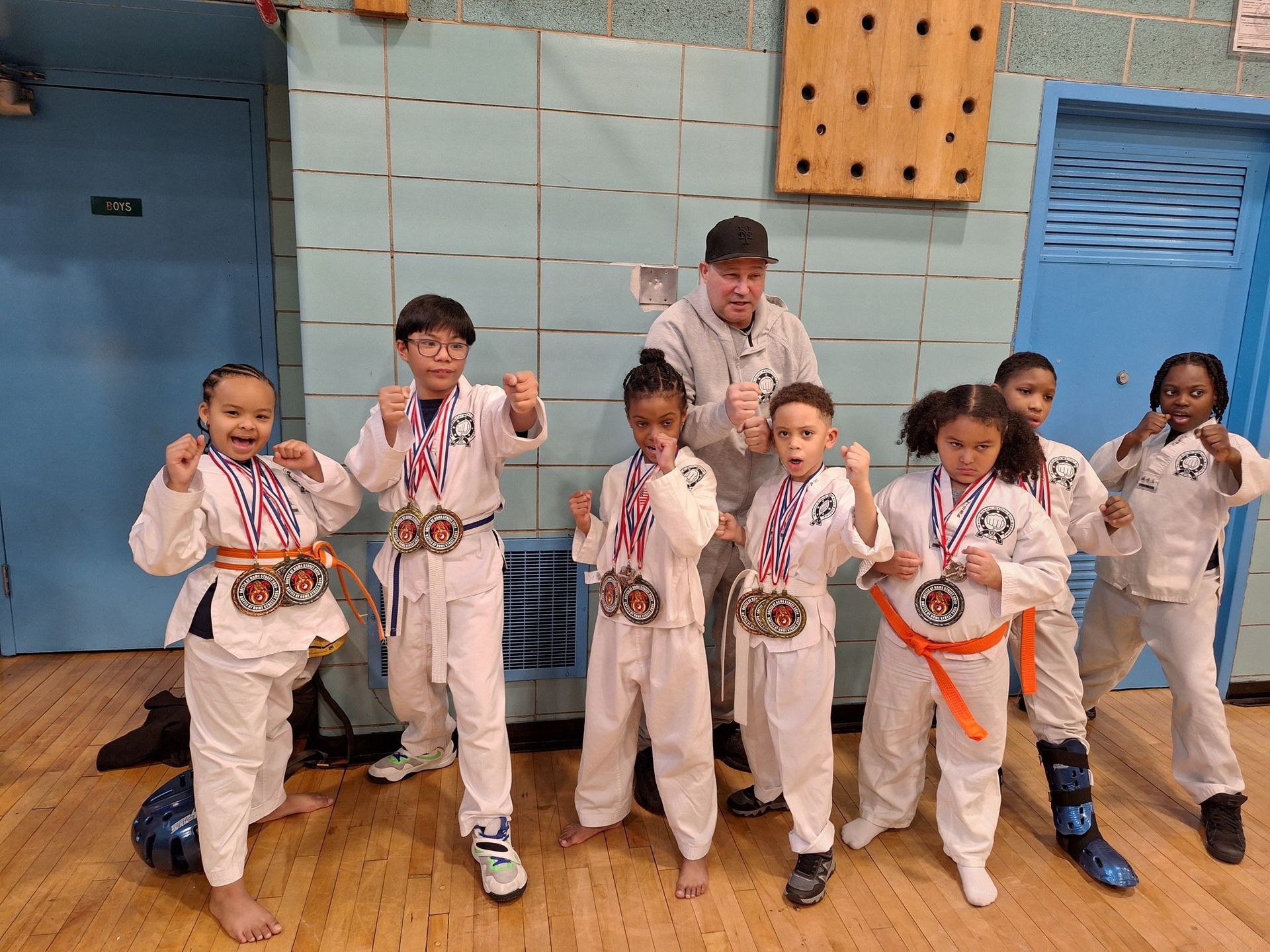 Group of kids in karate uniforms with medals, fists raised, posing with a coach in a gymnasium.