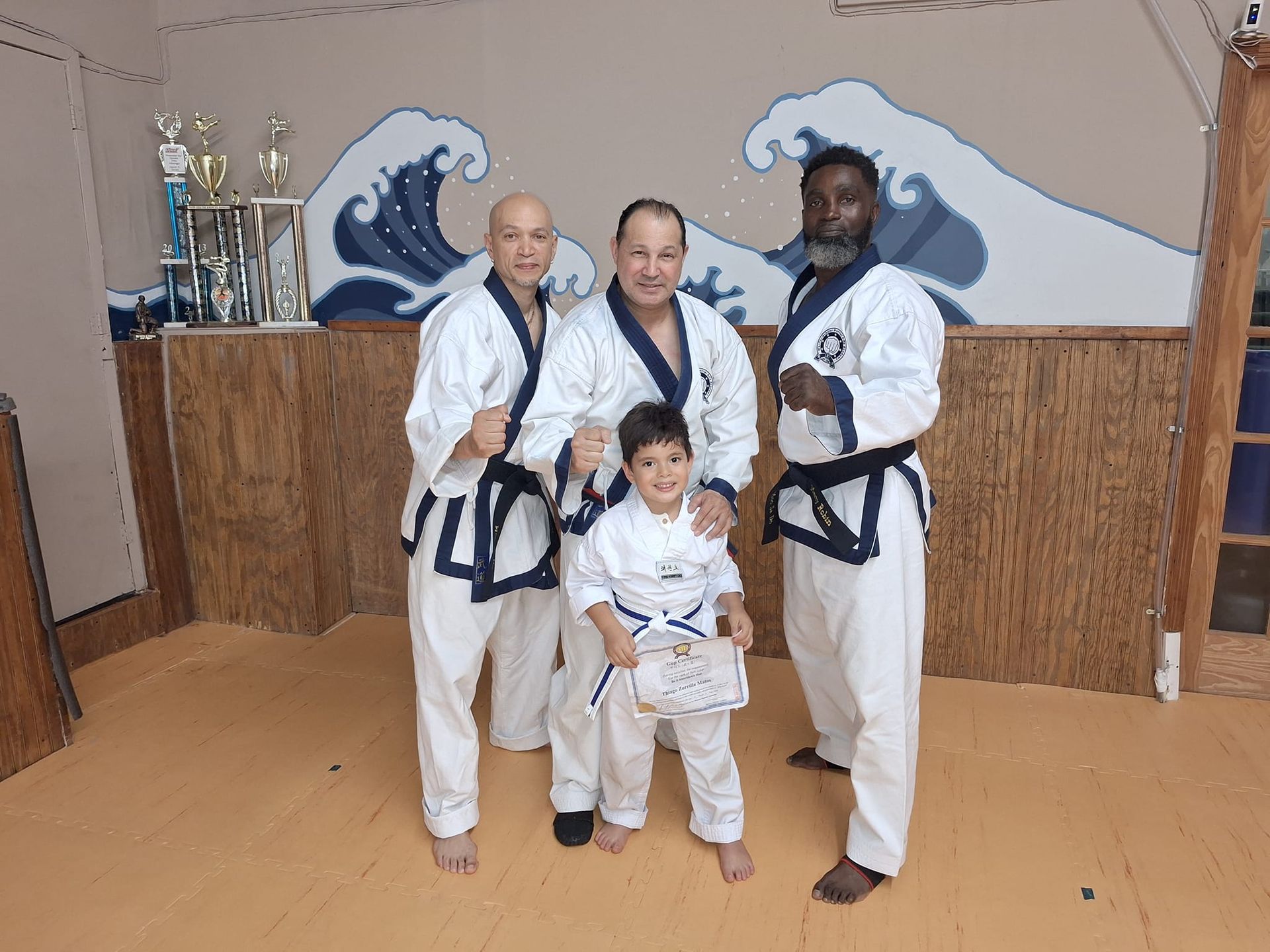 Four people in martial arts uniforms posing. Child holds certificate, adults smile in front of wave mural.