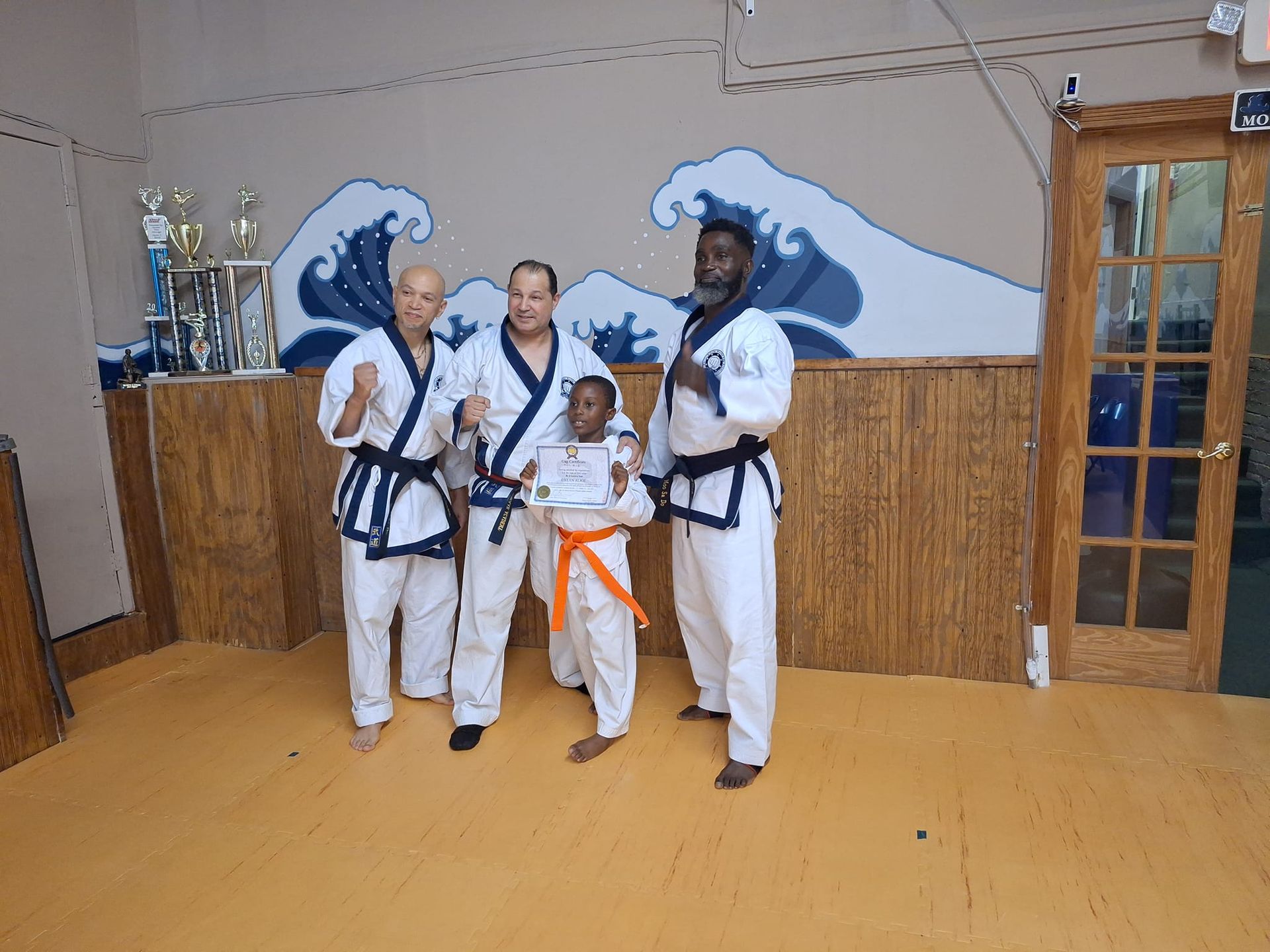 Group of martial artists in white uniforms, posing with an orange belt holder, inside a dojo with wave mural.