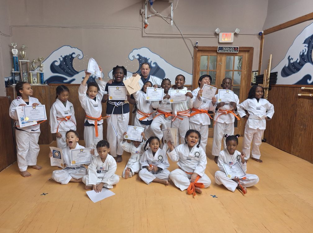 Group of young martial arts students in white uniforms, some holding certificates and smiling, in a dojo.