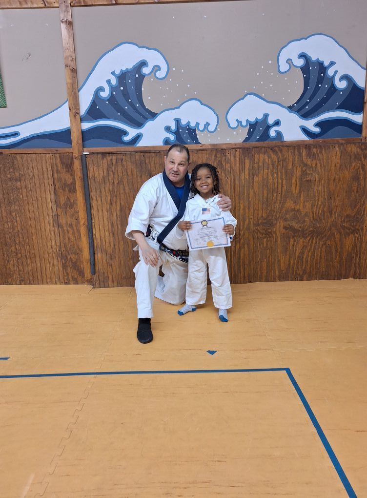 Man in karate uniform kneels with child in karate uniform holding a certificate in front of wave mural.