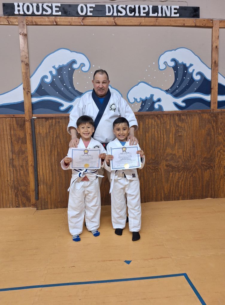 Two young boys in white karate uniforms, holding certificates, stand with an adult in a dojo.