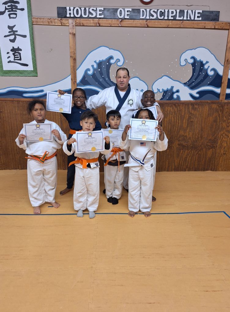 Martial arts class: Children in white uniforms with orange belts, holding certificates, pose with instructor.