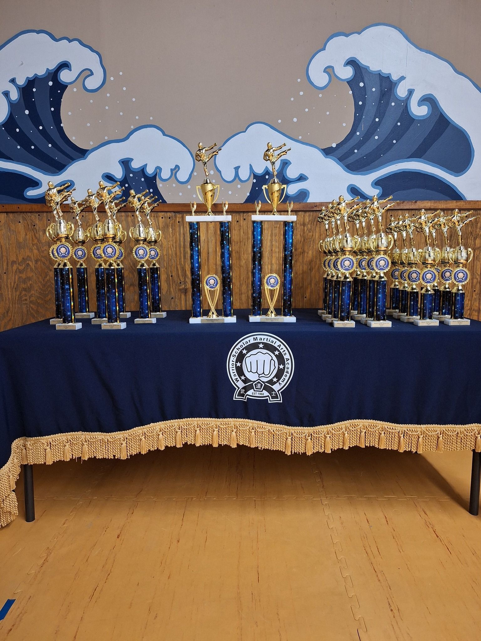 Trophies displayed on a blue table with a gold fringe, against a pixelated wave mural backdrop.