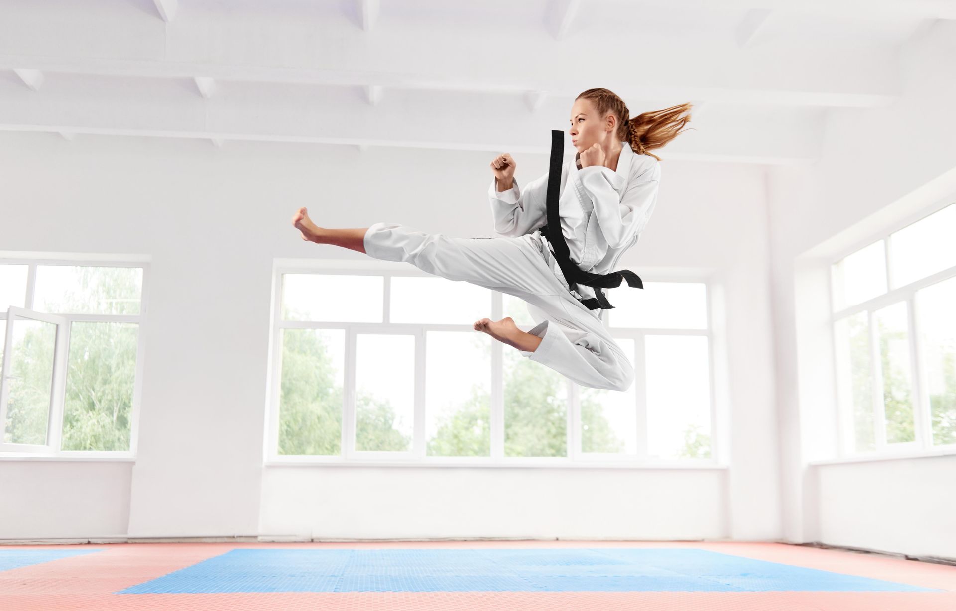Woman in karate uniform kicks high in the air; training room with windows.