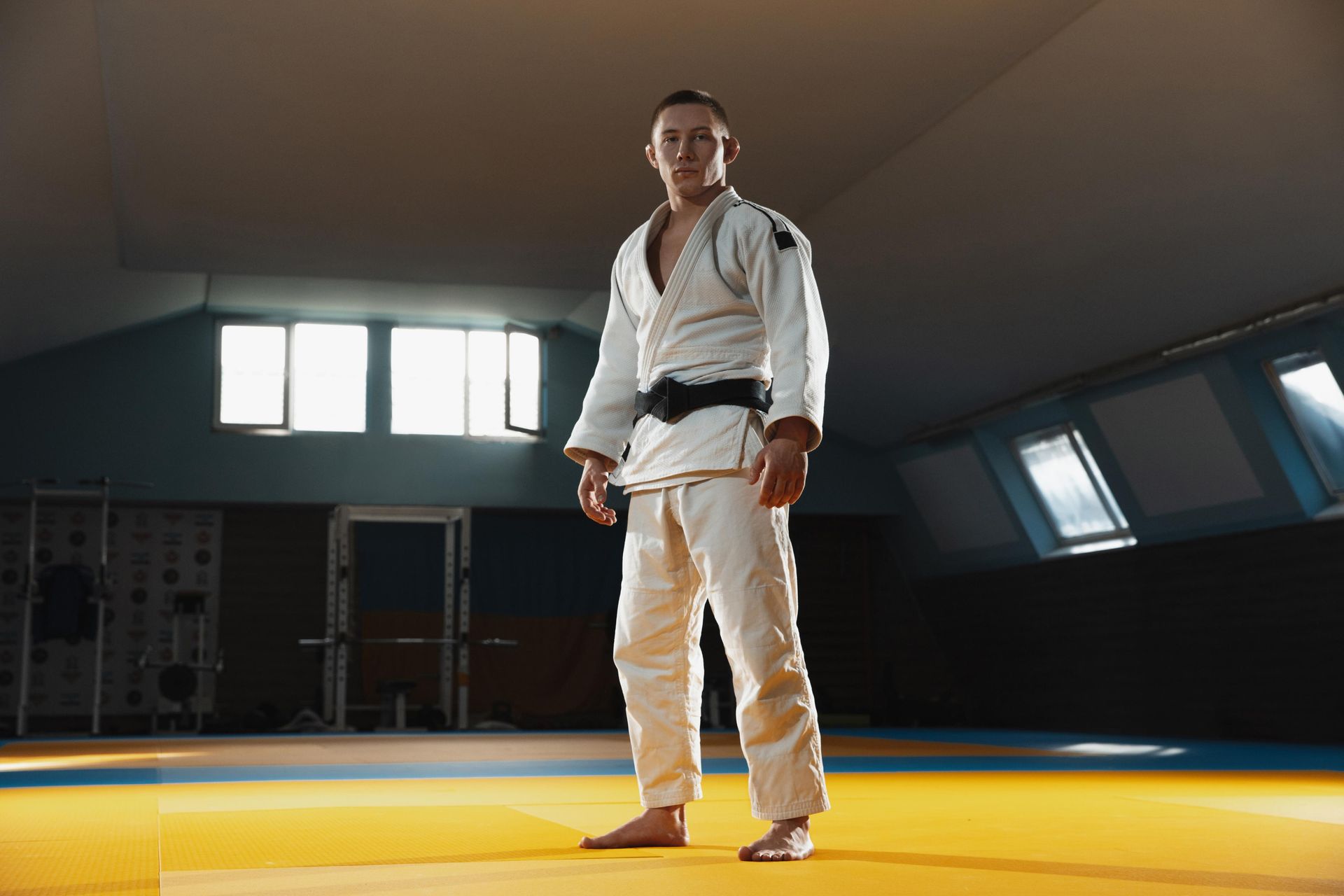 A judo athlete in a white uniform with a black belt, standing in a dojo with a focused expression.