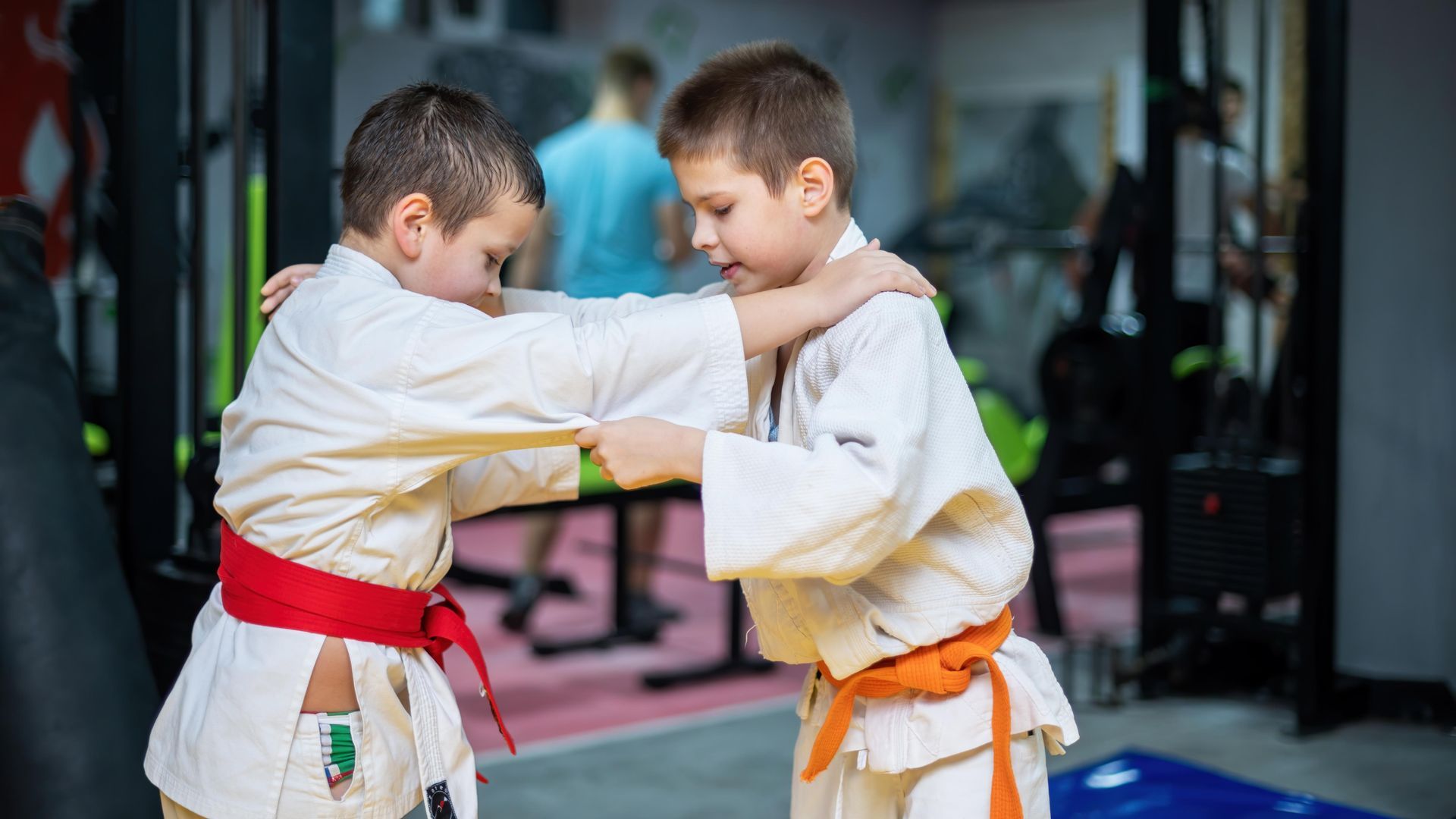 Two young boys in judo uniforms practicing in a gym. One wears a red belt, the other orange.