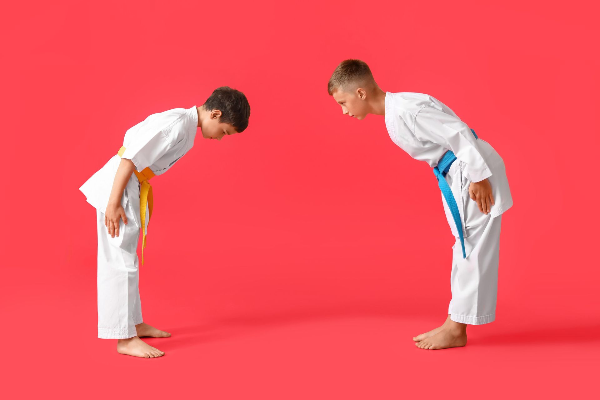 Two young karate students in white gis with colored belts bowing, red background.