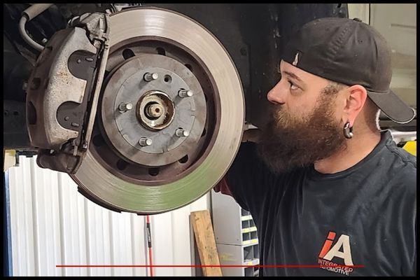 Mechanic examining a car's brake disc and caliper. He wears a hat and has a beard. | Integrated Automotive
