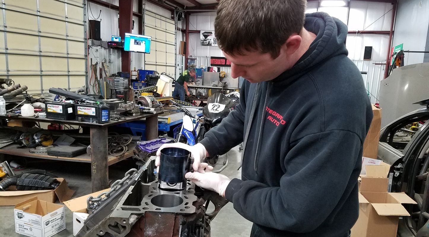 A mechanic in a black hoodie wearing gloves installs a piston into an engine block in a cluttered workshop. | Integrated Automotive