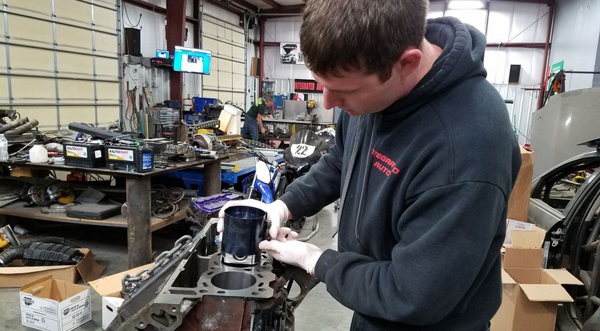 A mechanic in a black hoodie wearing gloves installs a piston into an engine block in a cluttered workshop. | Integrated Automotive
