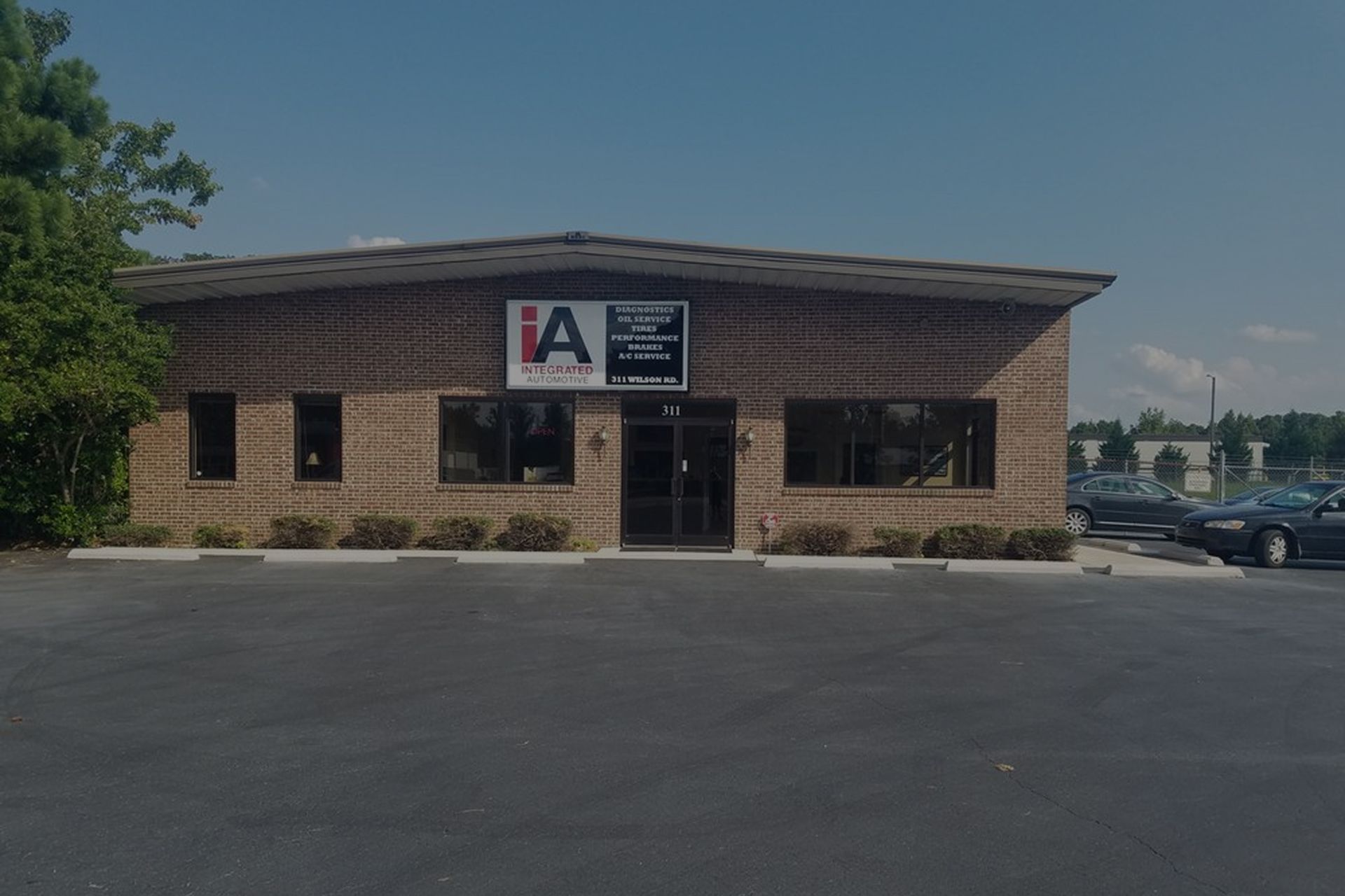 Brick building with IA logo over entrance, windows, and parked cars in the parking lot. | Integrated Automotive