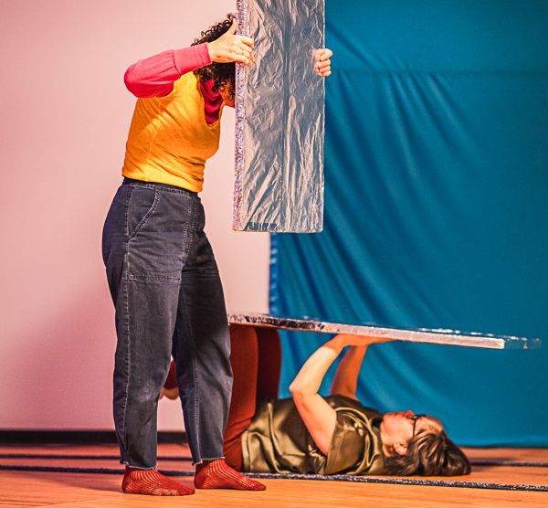 Two people manipulate silver, rectangular panels; one stands holding a vertical piece, the other lies below it.