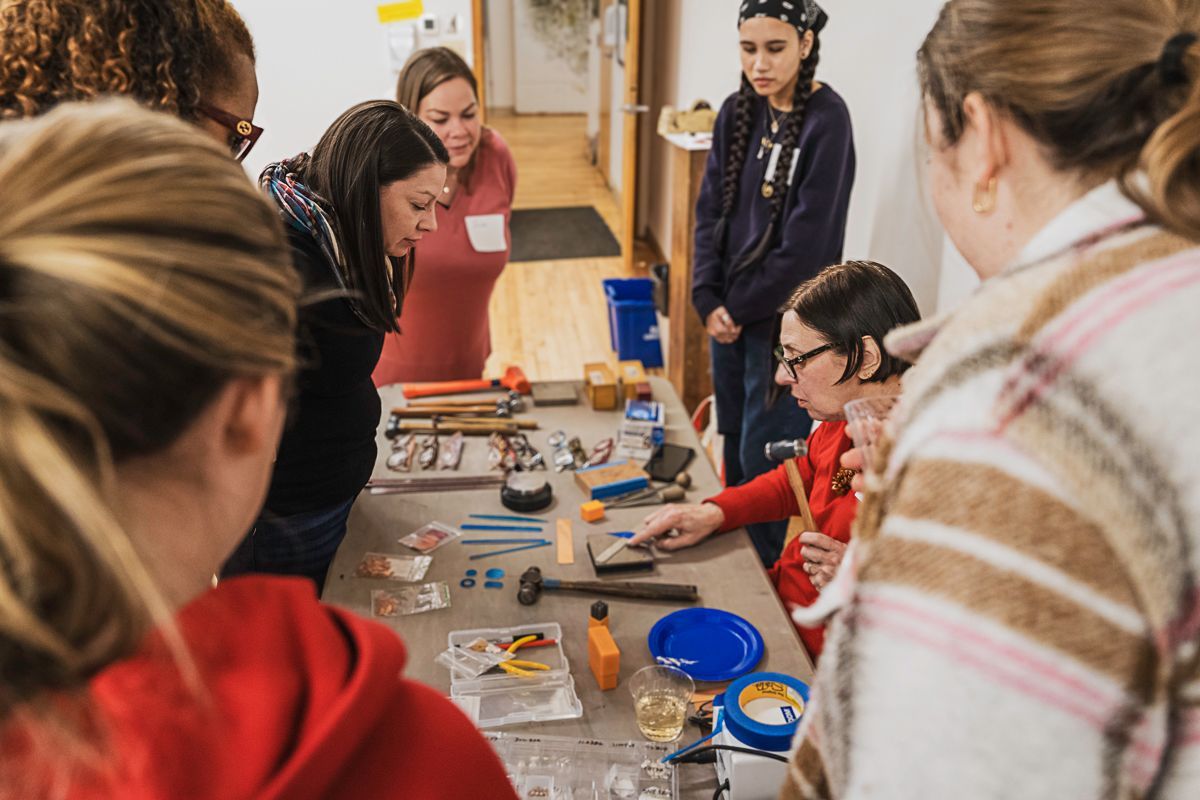 A group of people gathers around a table to observe a person demonstrating jewelry making with tools and supplies.
