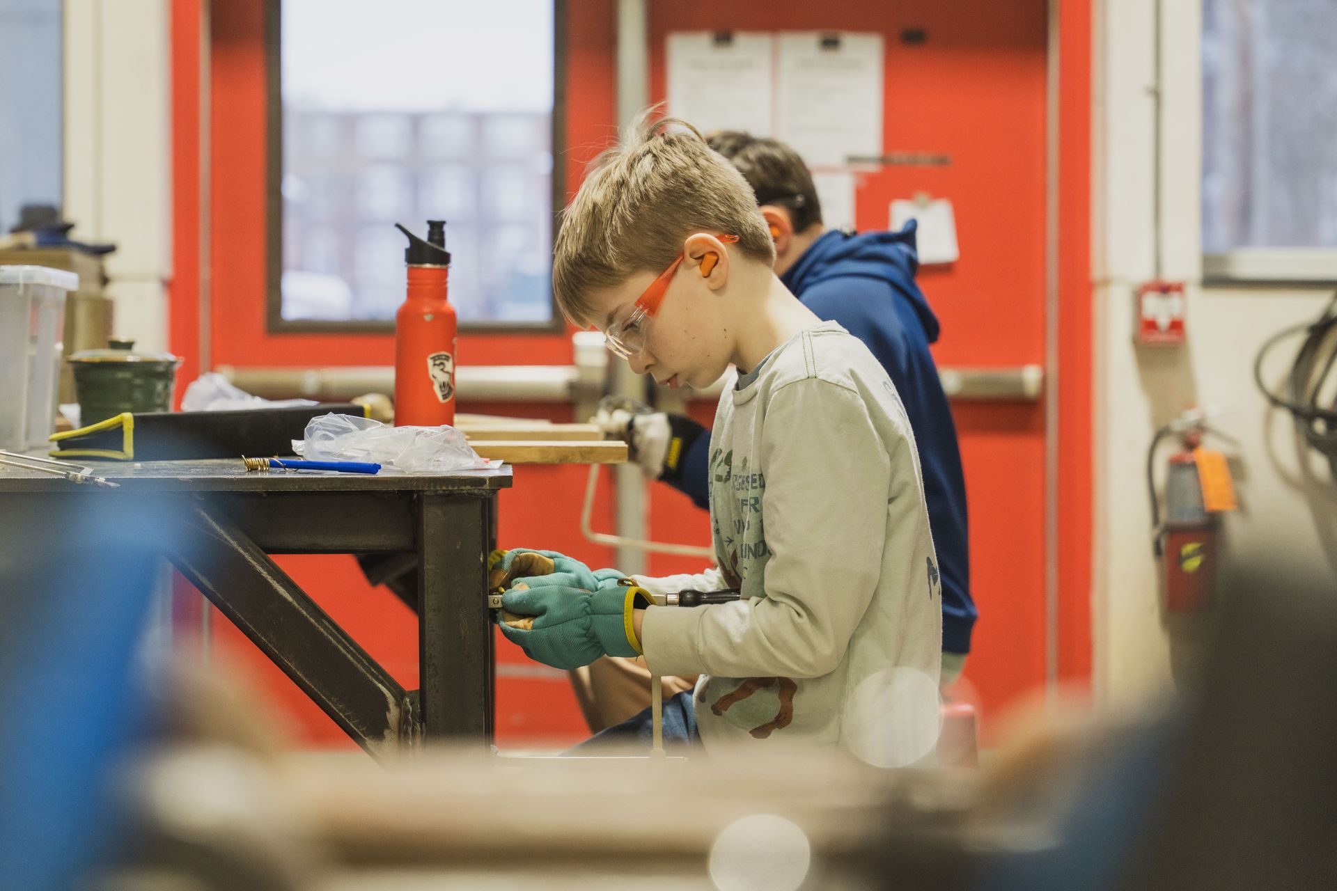 A person wearing safety goggles and work gloves uses a tool on a wooden project at a workshop table.