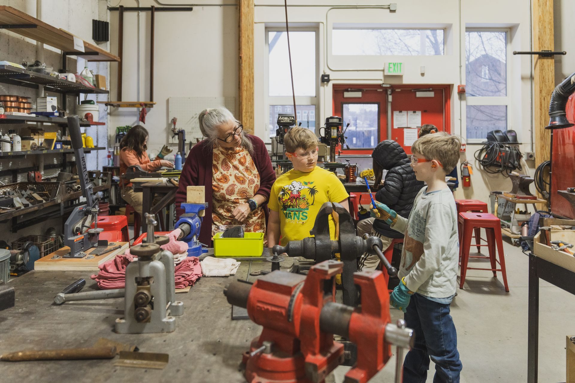 A workshop setting with people working at benches with tools, vises, and equipment, including a red door in the back.