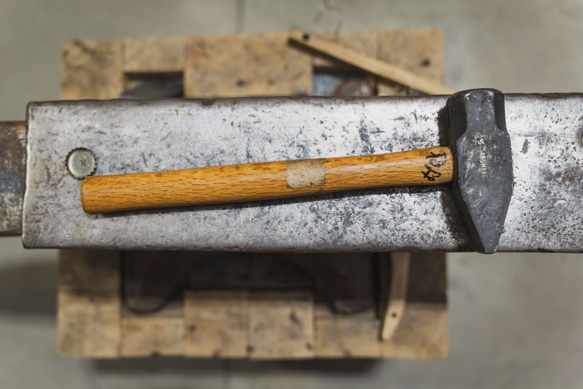 A metal hammer with a wooden handle rests on a heavy metal anvil set upon a wooden block.