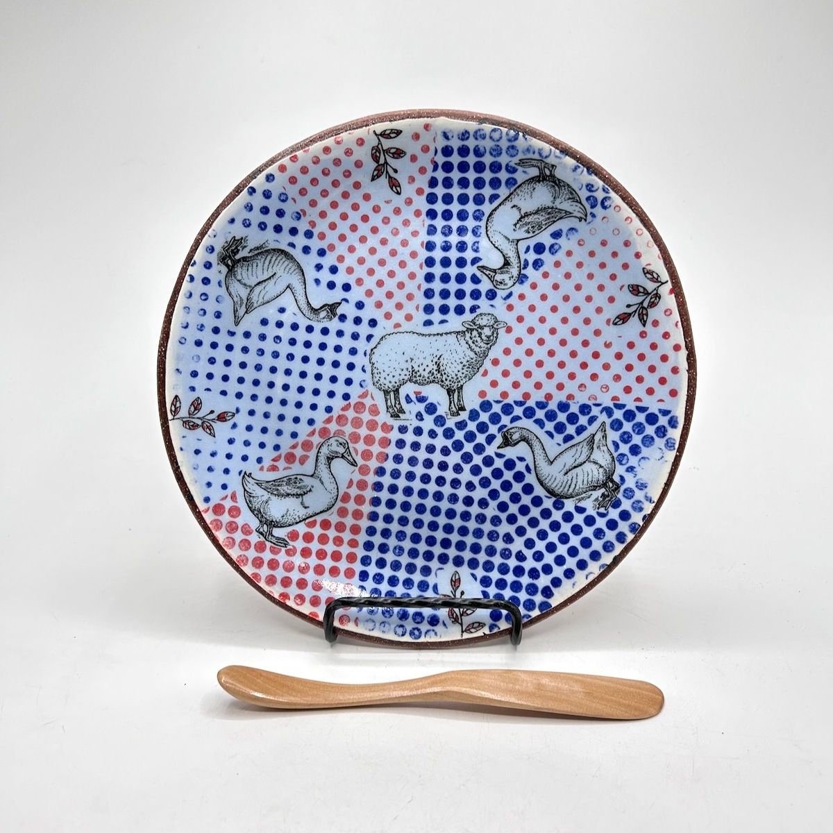 A decorative ceramic plate features a sheep and geese on a red and blue polka-dot background, with a small wooden spoon.