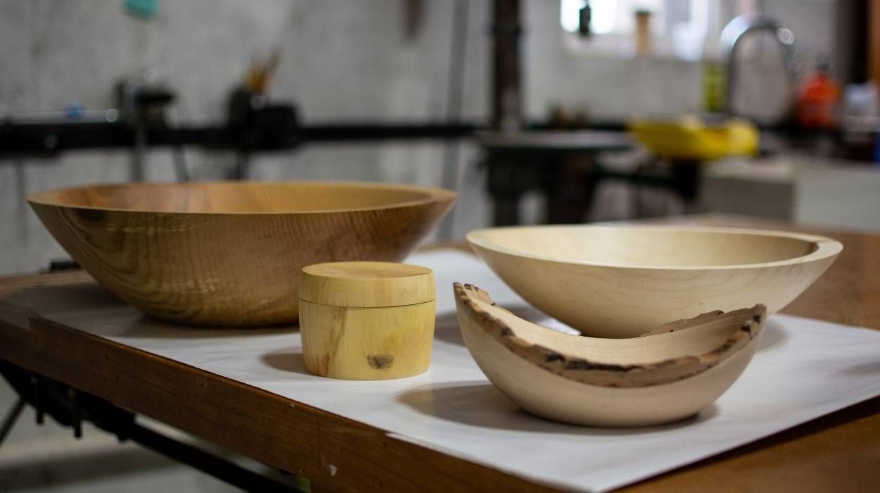 Several wooden bowls and a small lidded container rest on a workbench in a woodworking shop.