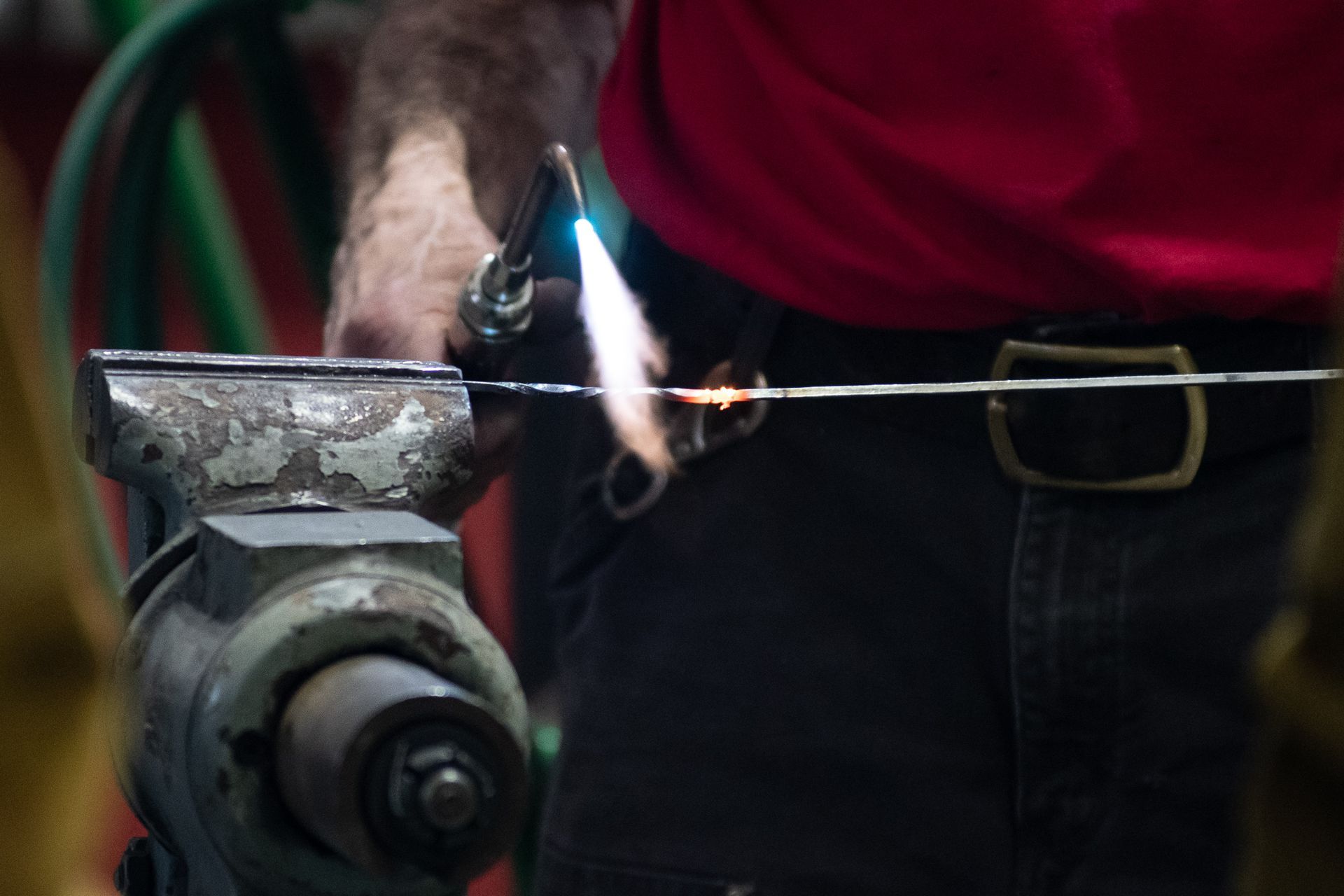 A person in a red shirt uses a blowtorch to heat a thin metal wire held in a metal vise.