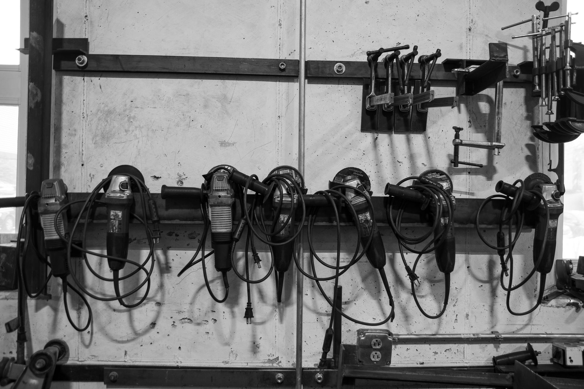 Black and white photo of a workshop wall with six angle grinders hanging on a rack below various metal clamps.