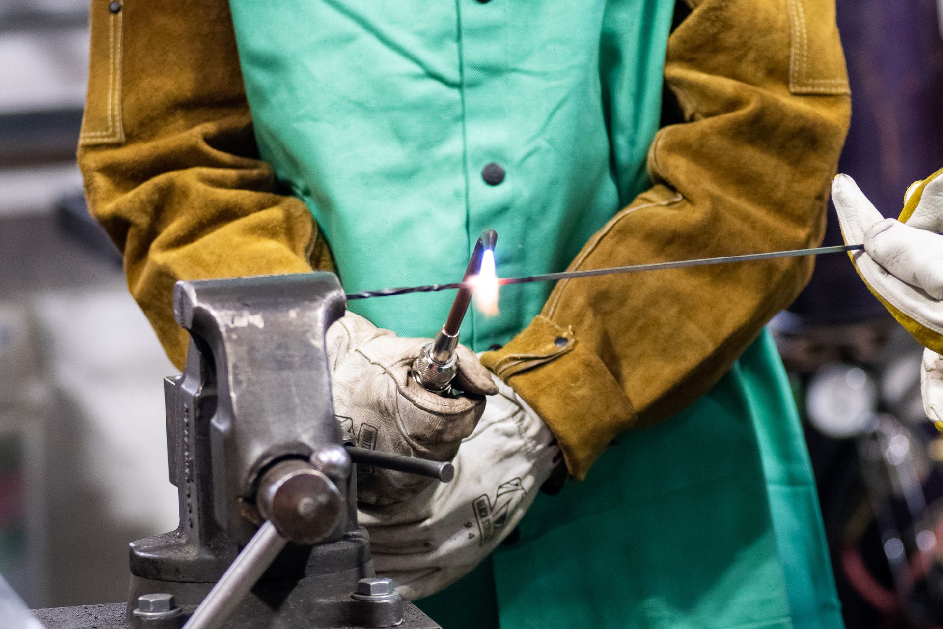 A person in a green and tan leather welding jacket uses a torch to heat twisted metal held in a bench vise.