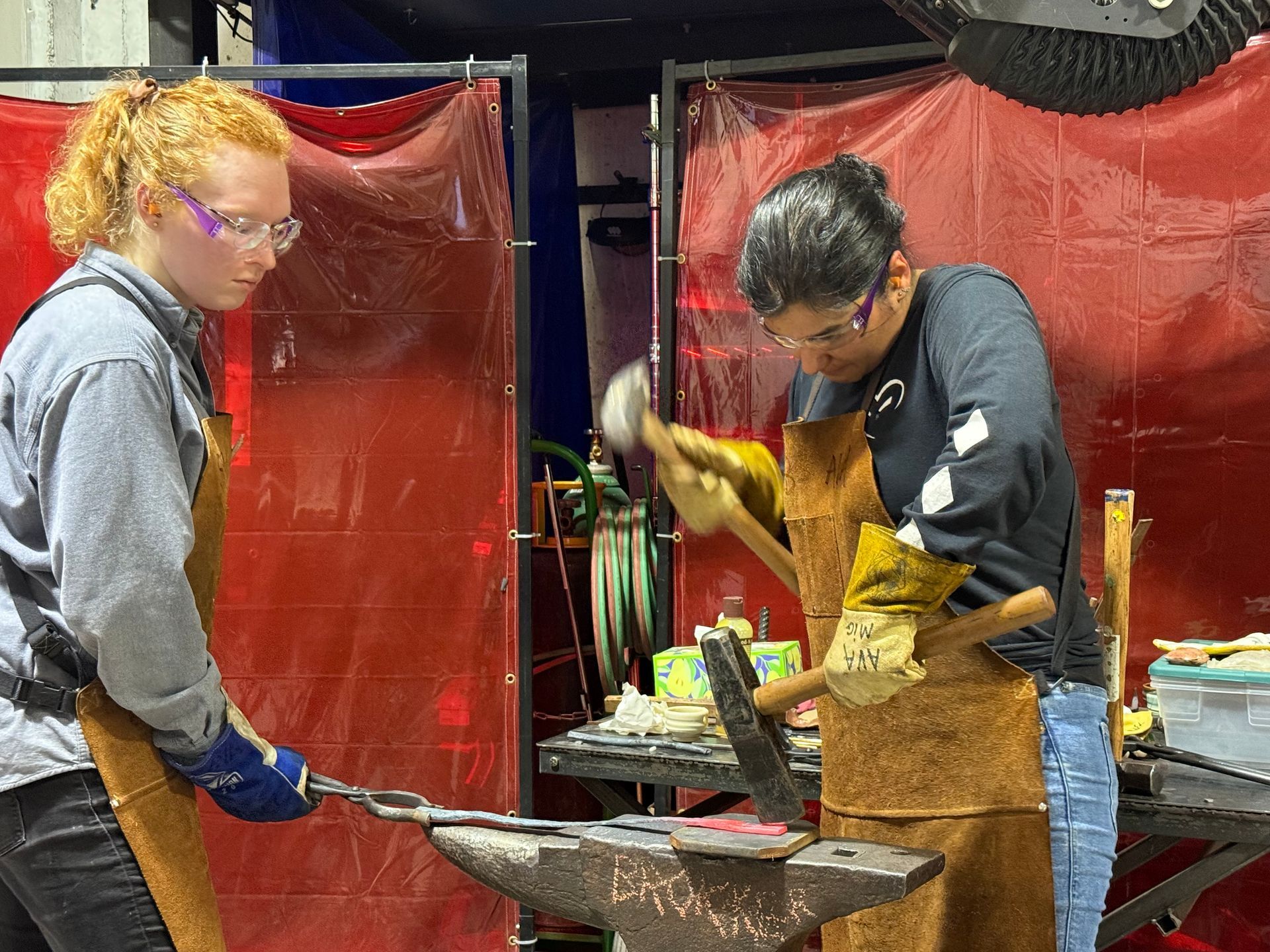 Two people in protective aprons use hammers and tongs to shape a piece of hot metal on an anvil in a workshop.