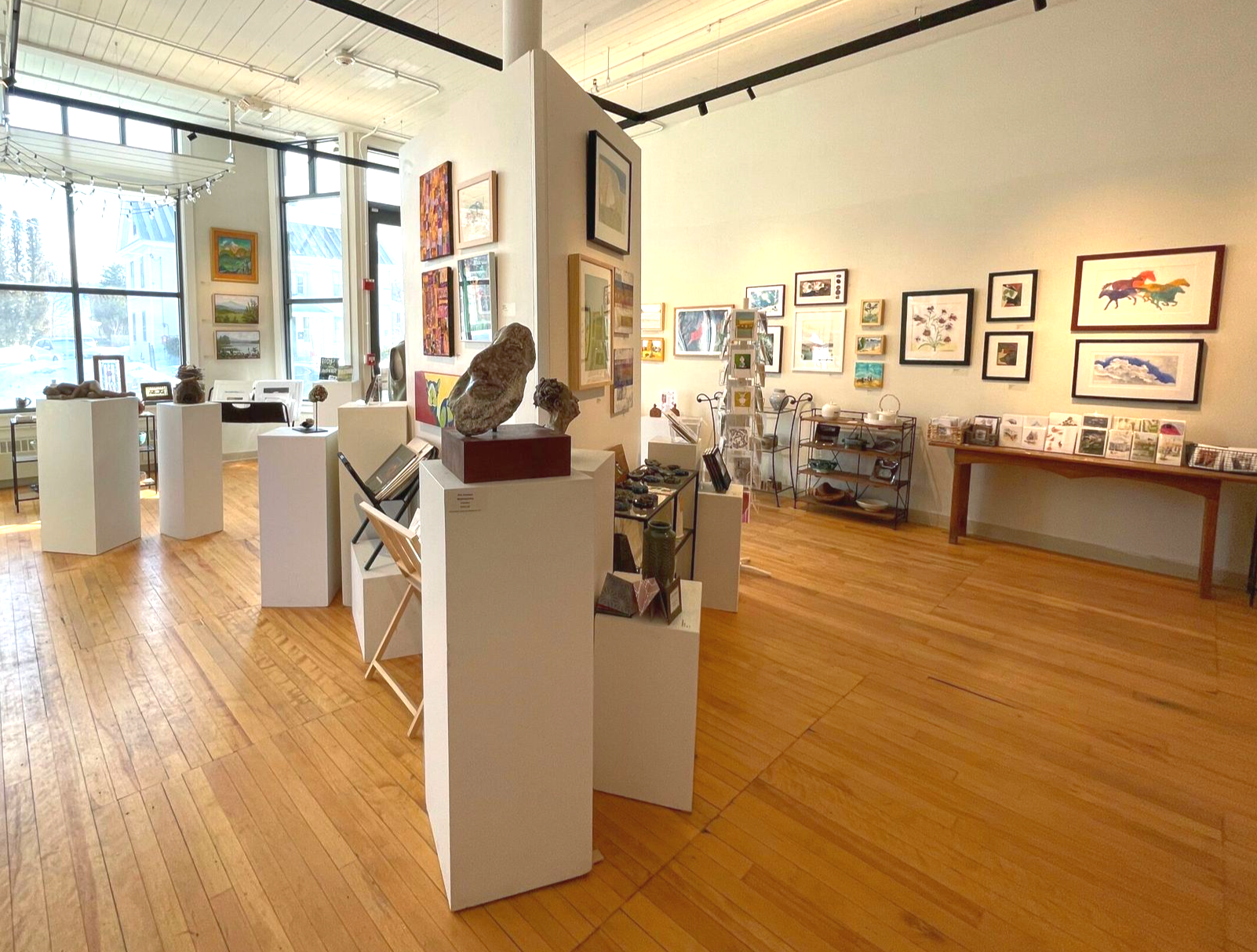 A bright art gallery with hardwood floors, display pedestals holding sculptures, and framed artworks on the walls.
