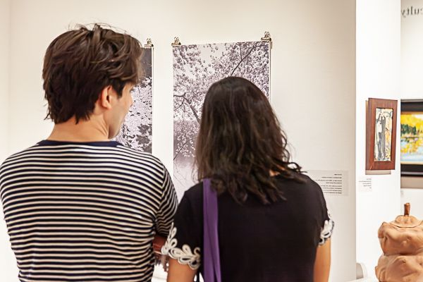 Two people viewed from behind examine two black-and-white landscape prints displayed on a white art gallery wall.