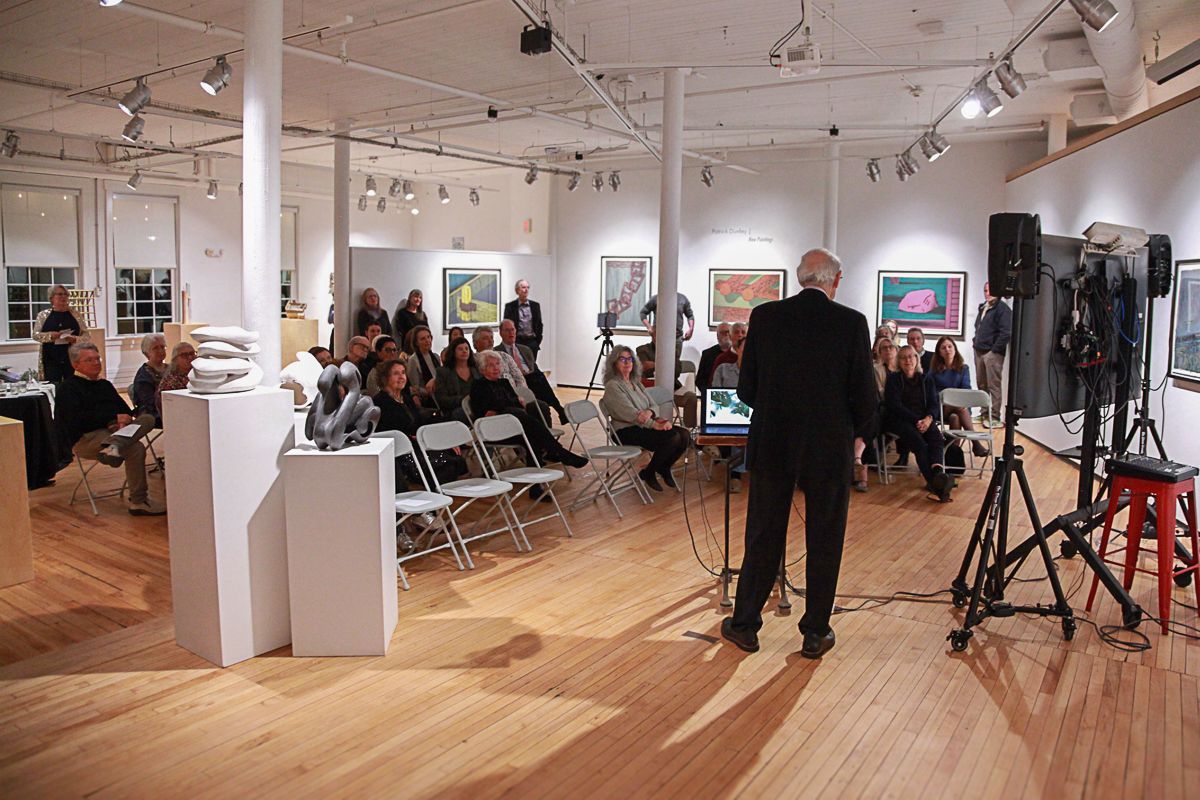 A presenter stands before an audience in a bright, wood-floored gallery with art displayed on the white walls.