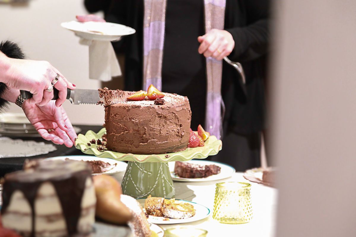 A person cuts a chocolate-frosted layer cake on a green pedestal stand, with another cake and dessert on a table nearby.