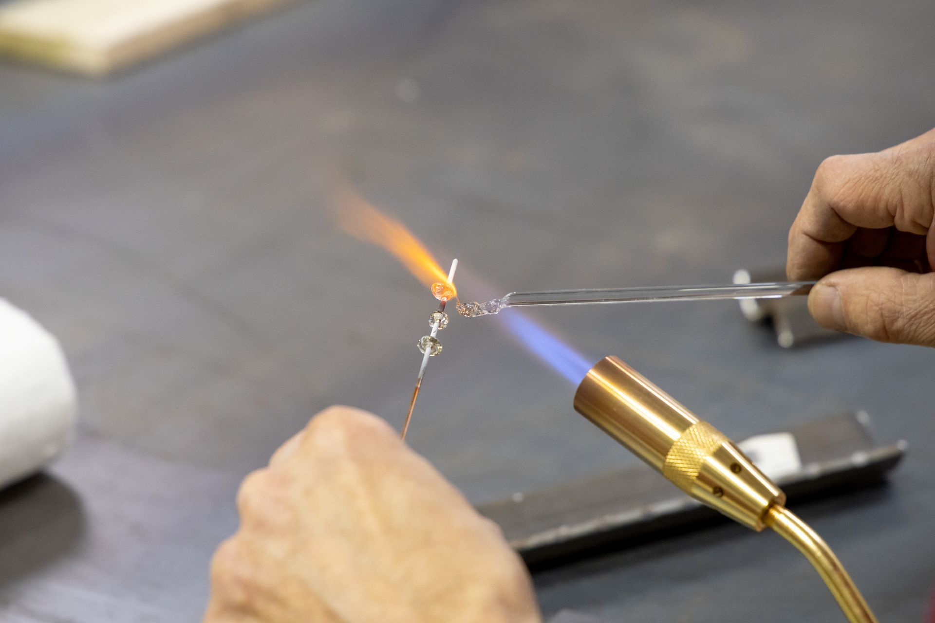 A person uses a torch to melt and shape glass on a metal rod over a workspace.