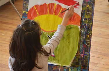 A person painting a red and yellow flower on a canvas on an easel, seen from behind.