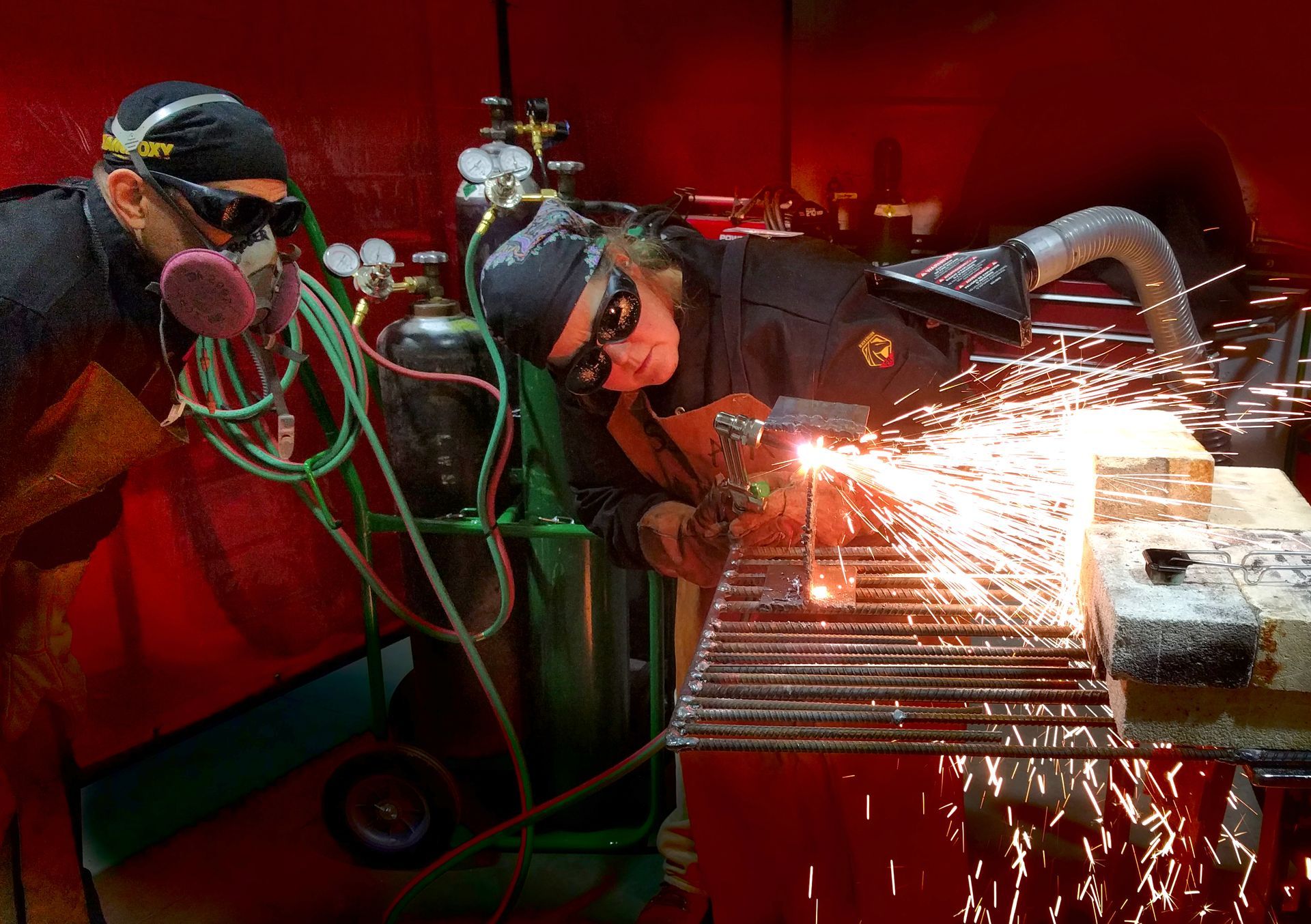 Two people in protective gear and goggles watch sparks fly as one uses a torch to cut metal at a workbench.