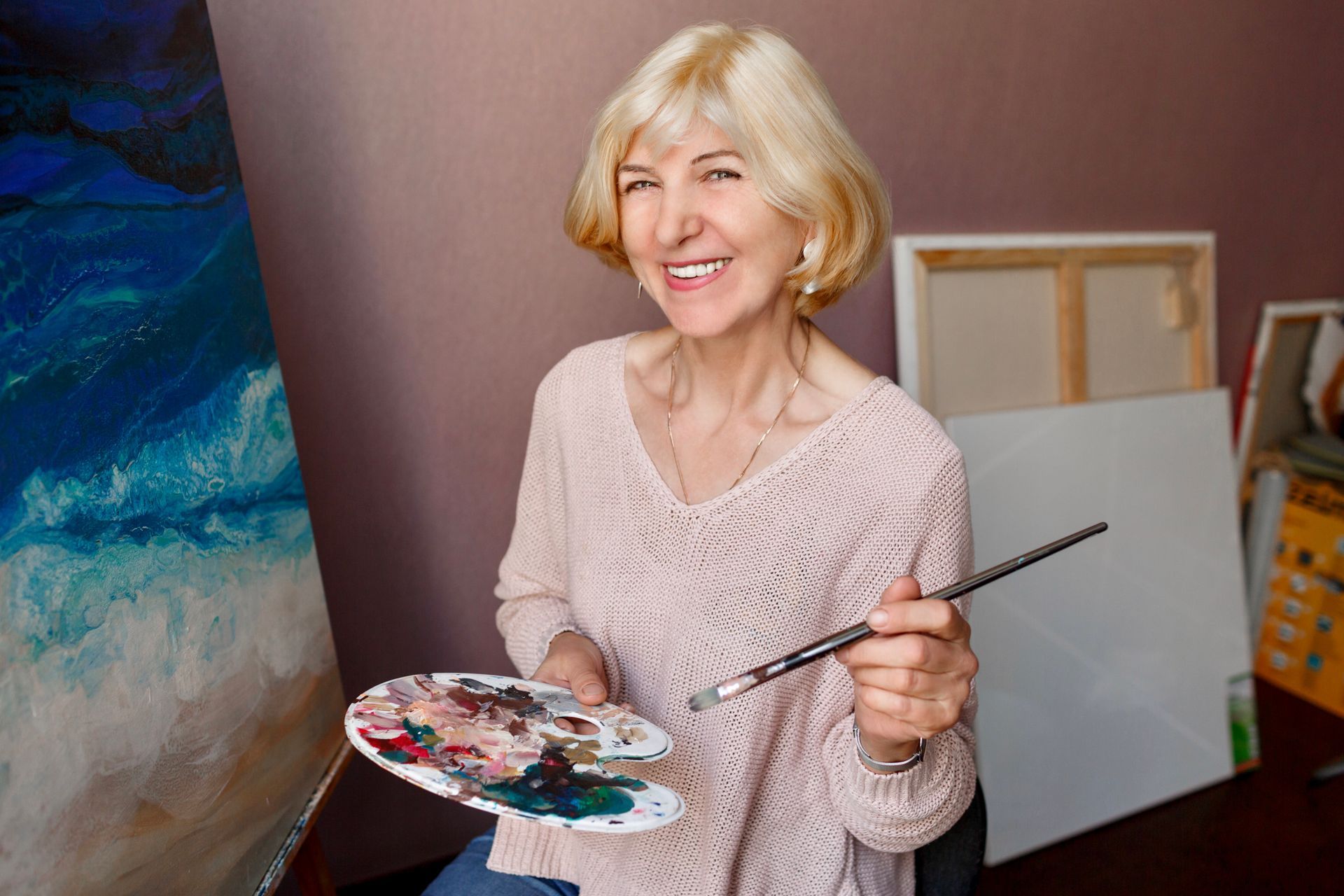 A smiling person holds a paint palette and brush in front of a blue abstract painting in an art studio.
