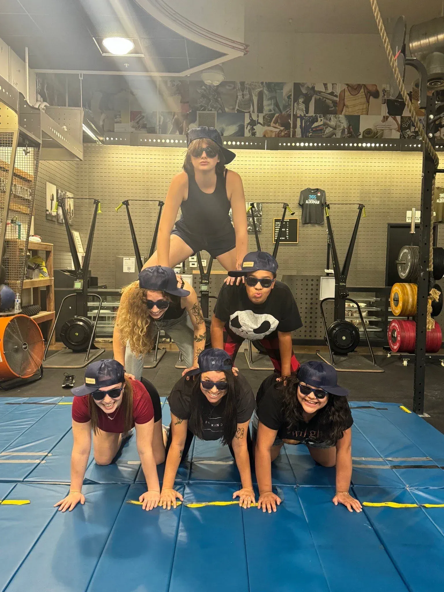 Six people in a human pyramid formation at a gym, all wearing sunglasses and caps.