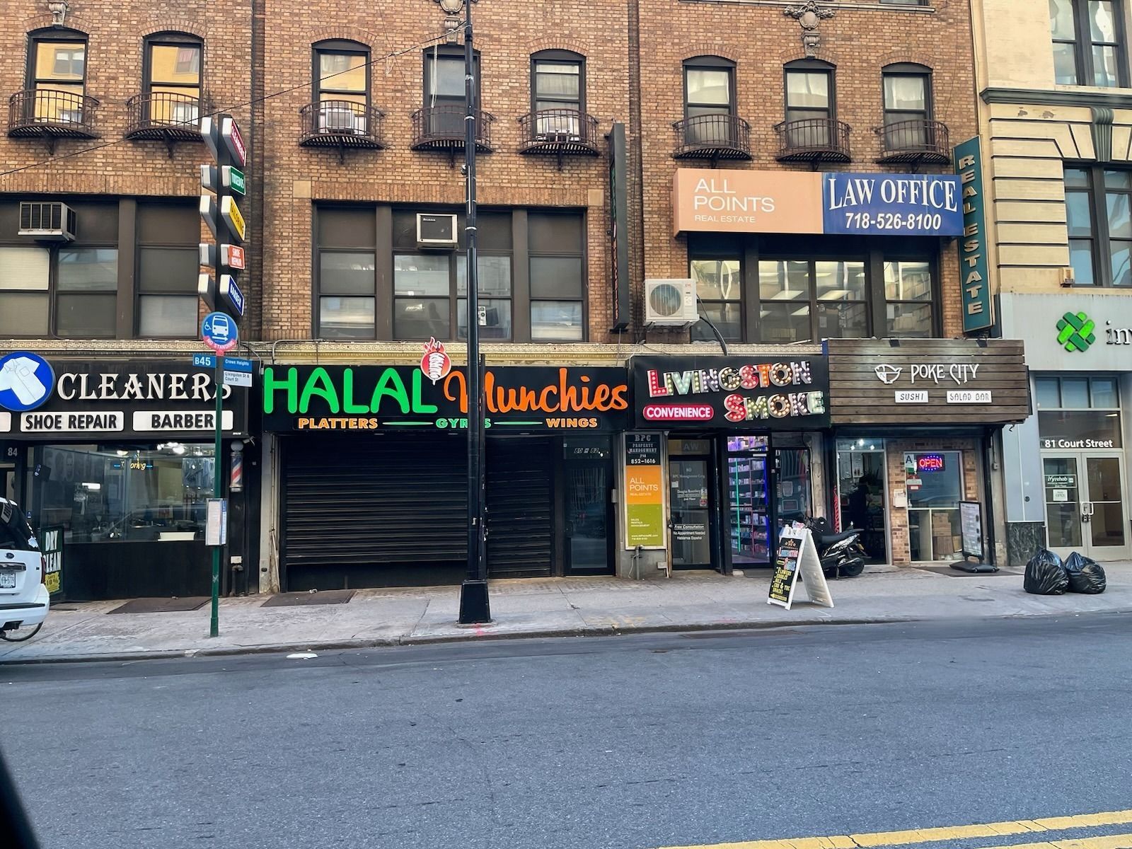 Street-level view of storefronts, including a cleaner, Halal restaurant, and smoke shop. A dog sits on the sidewalk.