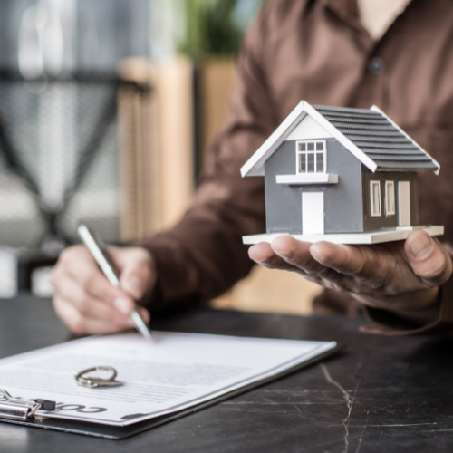 A person is writing on a clipboard while holding a model house