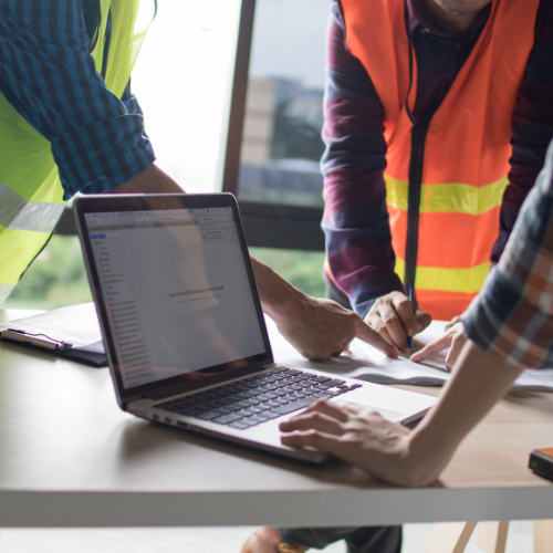 Two men in safety vests are working on a laptop