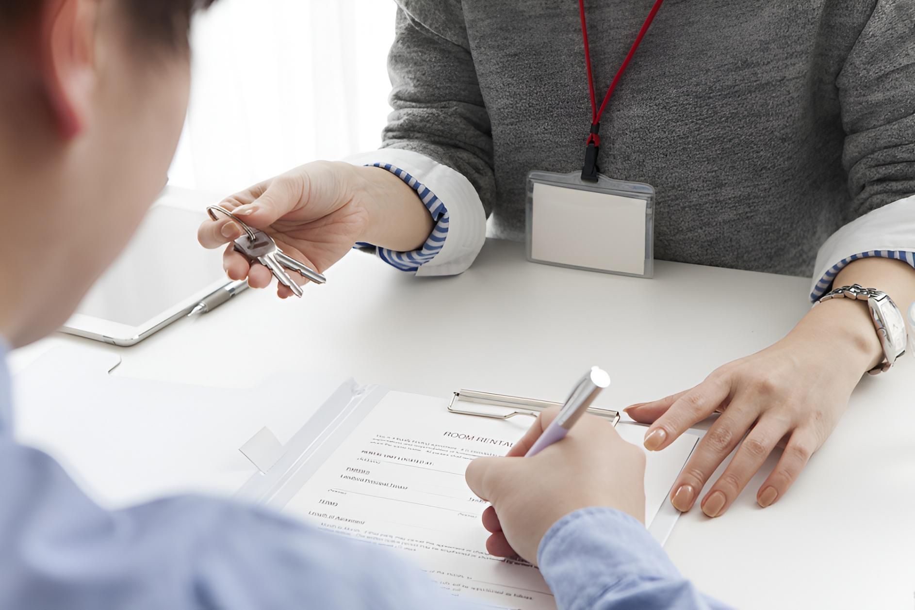 A Person Signing Paperwork at a Desk While Another Person Holds Out Keys — Morris Legal & Conveyancing in Forster, NSW