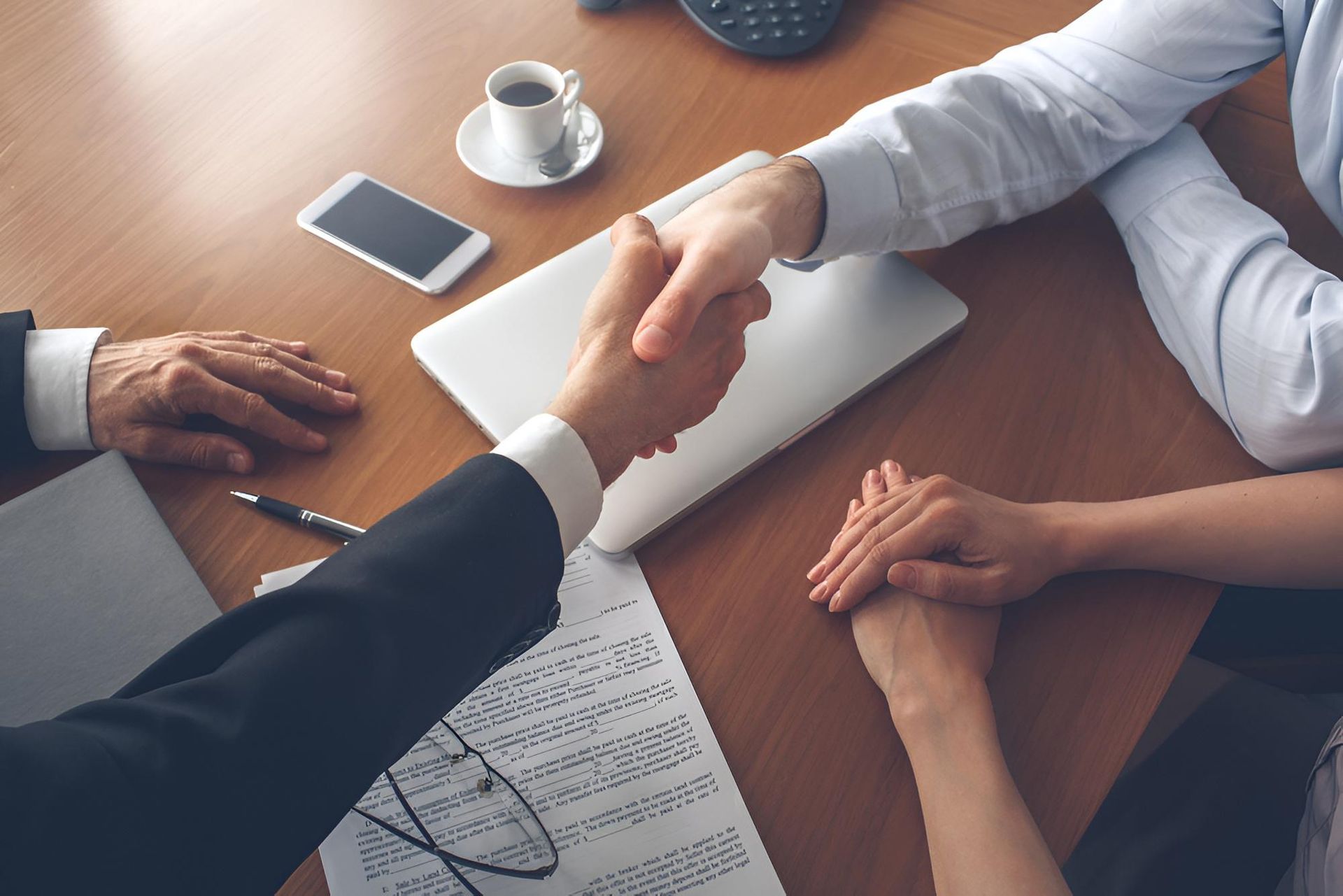 Two People in Business Suits Shaking Hands Over a Wooden Table, Laptop, Papers, and Coffee Cup Present — Morris Legal & Conveyancing in Forster, NSW