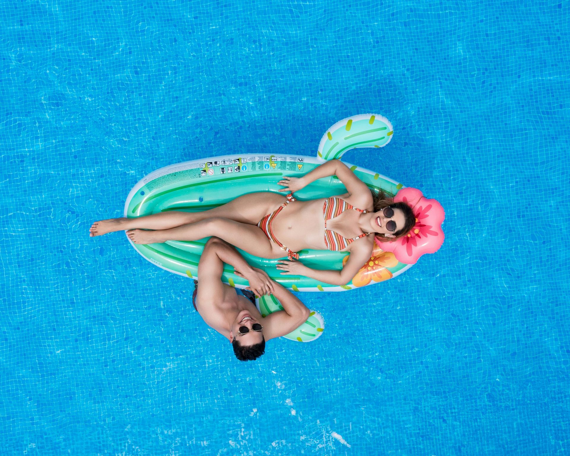 Couple relaxing on a cactus float in a blue pool.