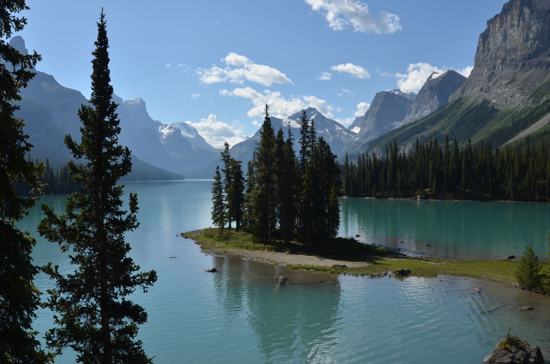 Een turquoise meer met een klein eilandje en altijdgroene bomen, bergen op de achtergrond onder een blauwe hemel.