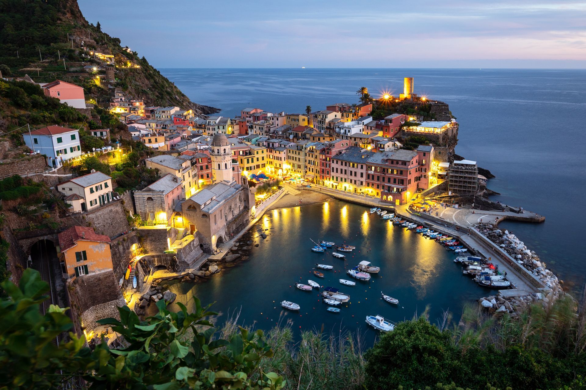 Een kustdorpje dat bij schemering verlicht is, kleurrijke gebouwen, boten in de haven en een kasteel op een heuveltop.