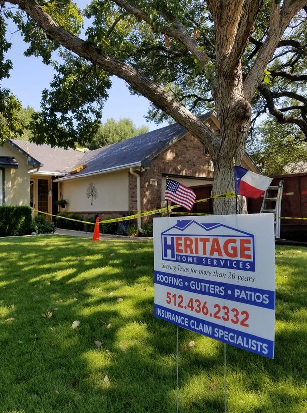 Sign for Heritage Home Services, roof being worked on, in front of a house, with flags and caution tape.