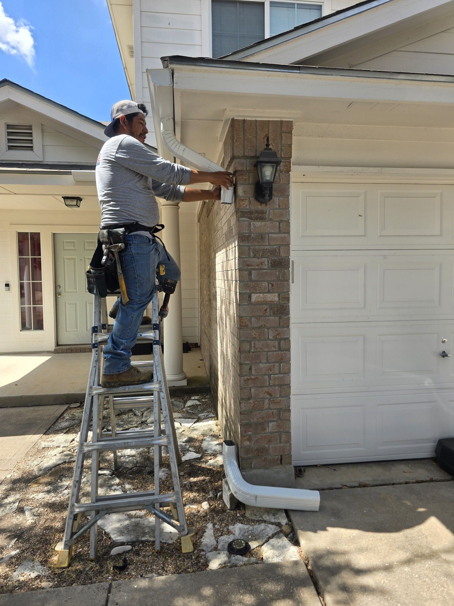 A person on a ladder repairs an exterior light fixture on a house near a garage.